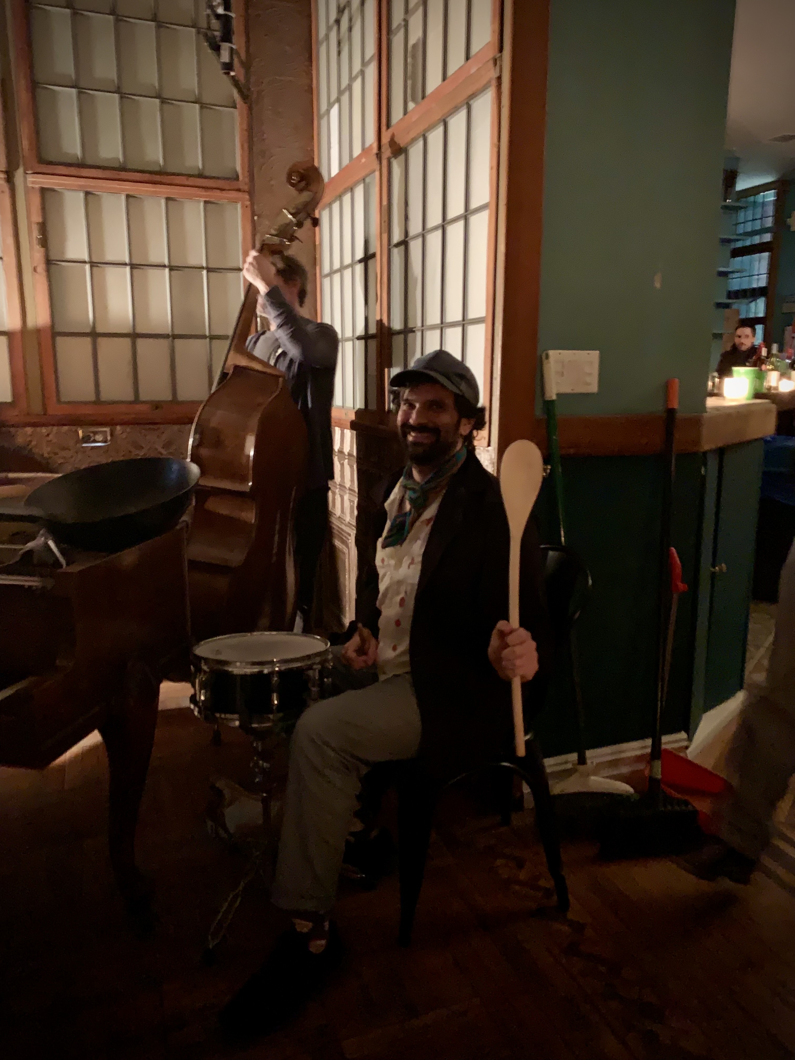 A smiling drummer holding a wooden spoon beside the piano.