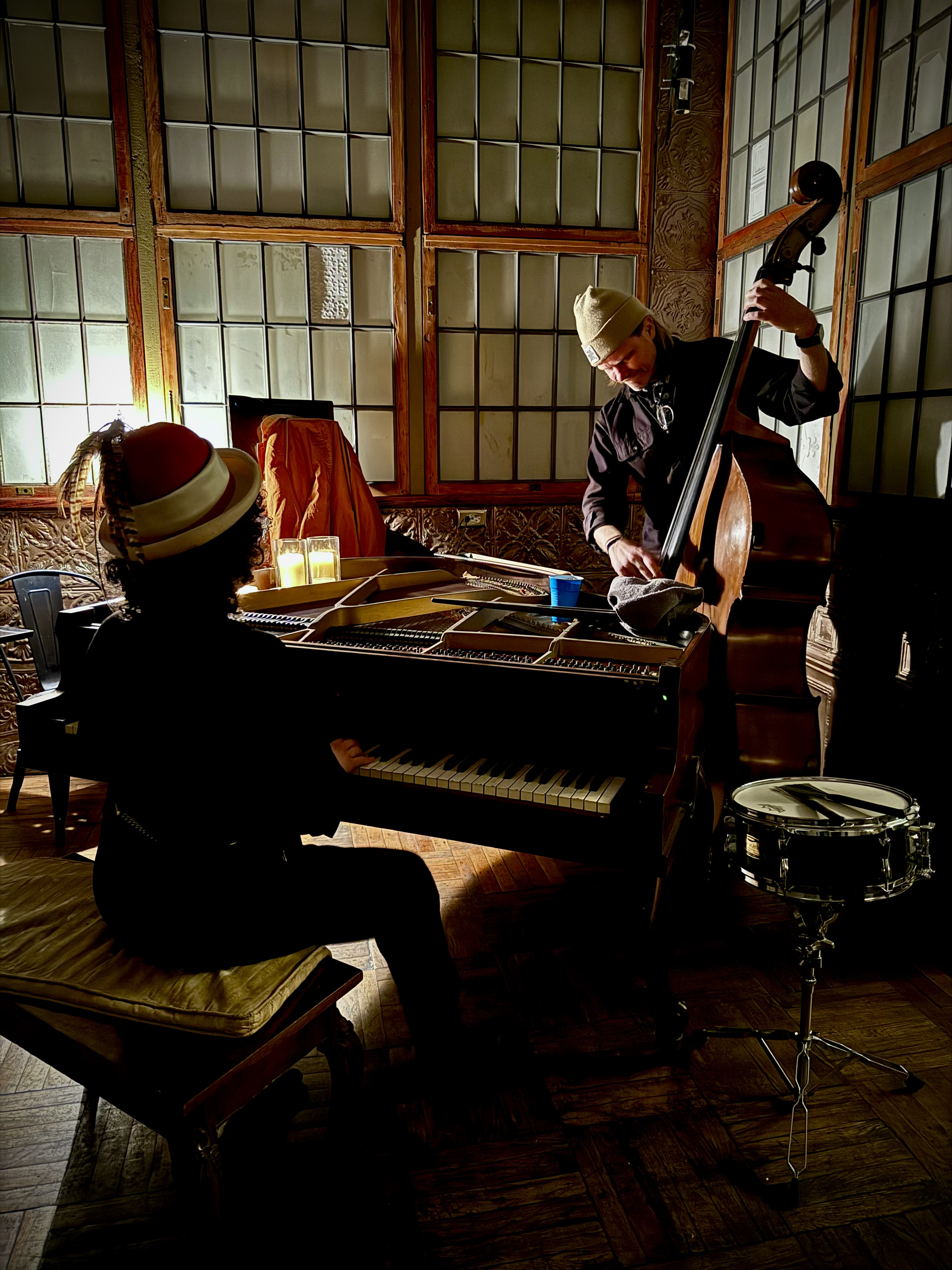 A pianist and bassist playing around the old piano in the candlelit room.